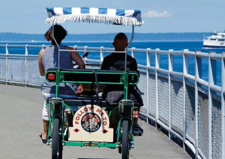 Two people ride a four-wheeled pedal cart along a waterfront path in the West Seattle neighborhood, shaded by a blue-and-white striped canopy. There is water, a distant boat, and a “Follow Me To” sign on the back of the cart.