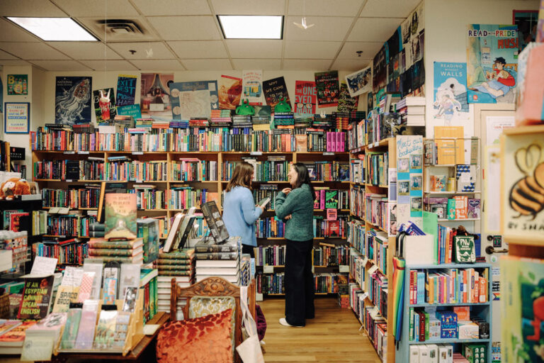 Two women stand and talk in the middle of a colorful, cozy bookstore on Bainbridge Island, surrounded by shelves of books, posters, and displays. Stacks of books and various book-themed decorations cover the space, creating a warm haven that attracts visitors from nearby Bremerton and beyond.