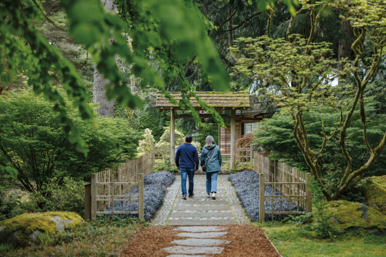 Two people walk hand in hand down a stone path through a lush green garden toward a wooden gate, surrounded by trees and foliage, evoking the tranquil charm of Bainbridge Island and the peaceful atmosphere found in Bremerton.