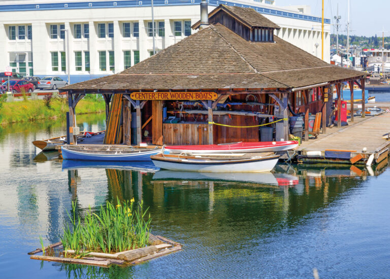 A wooden boathouse labeled "Center for Wooden Boats" sits on a dock in the South Lake Union neighborhood, surrounded by small wooden boats and calm water, with a building and cars in the background.