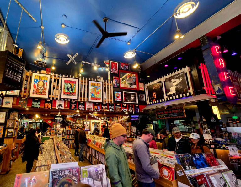 People browse vinyl records in a colorful, crowded record store nestled in the heart of the West Seattle neighborhood, with posters and album covers on the walls, under bright lights and a blue ceiling. A neon "CAFE" sign glows on the right.