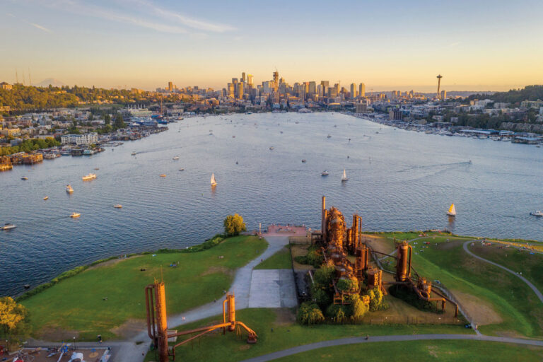 Aerial view of Gas Works Park with industrial structures in the foreground, nestled between the Fremont & Wallingford neighborhoods, boats on Lake Union, and the Seattle skyline including the Space Needle in the background at sunset.