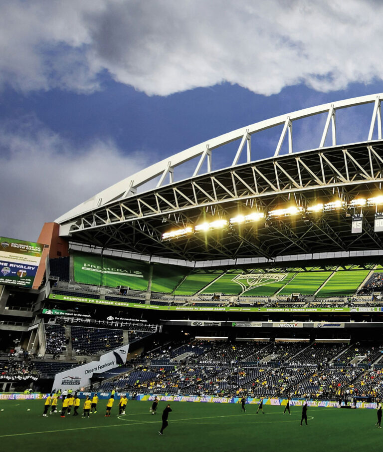 A soccer stadium in the heart of the SoDo and Georgetown Seattle neighborhoods, with a partially filled crowd, players warming up on the field, and a dramatic cloudy sky overhead. Bright stadium lights illuminate the seating area and green pitch.