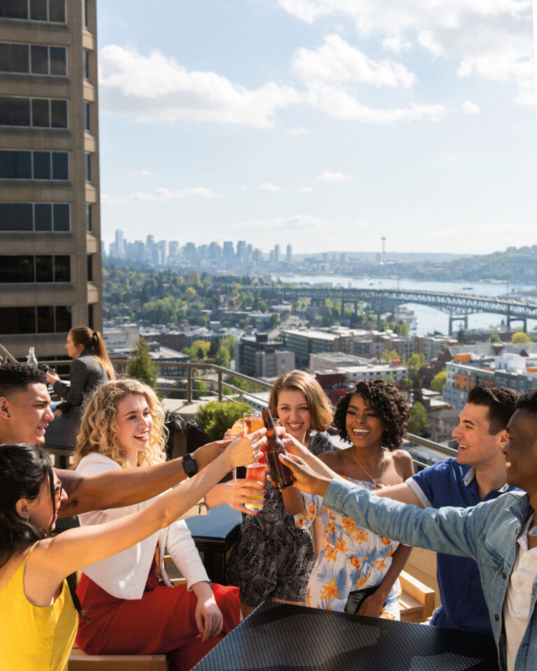 A group of friends sits at an outdoor table in the lively University District Seattle neighborhood, raising drinks in a toast with a city skyline and Space Needle in the background on a sunny day.