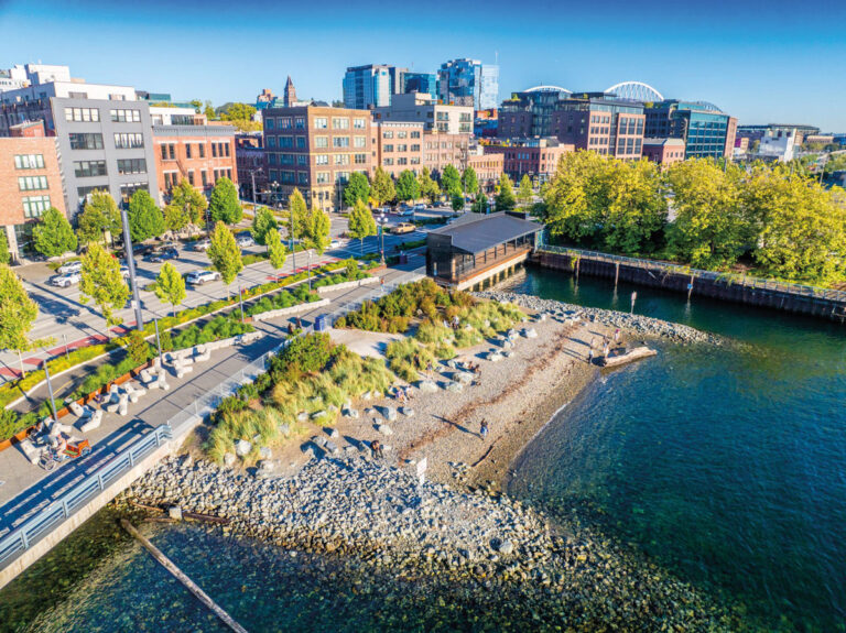 Aerial view of an urban waterfront park in the Pioneer Square neighborhood, with trees, a rocky shoreline, people walking, and city buildings in the background under a clear blue sky.
