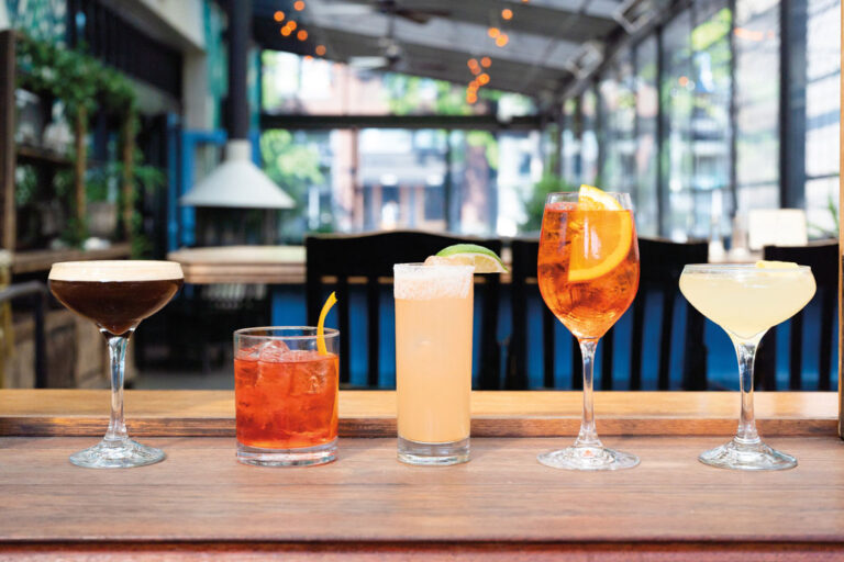 Five colorful cocktails in different glasses are lined up on a wooden bar counter in a bright, modern indoor-outdoor restaurant setting in the lively Ballard neighborhood, with plants and string lights glowing in the background.