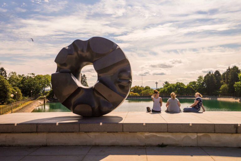 A large abstract sculpture sits beside a reflecting pool in the Capitol Hill neighborhood, with three people on the edge enjoying a sunny, partly cloudy day. Trees and greenery surround the water in the background.