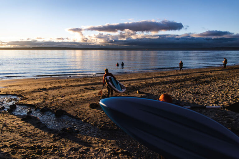 People on a sandy beach at sunset in the Ballard neighborhood, some near the water and one person carrying a surfboard. Another surfboard is visible in the foreground, with calm waves and clouds over the horizon.