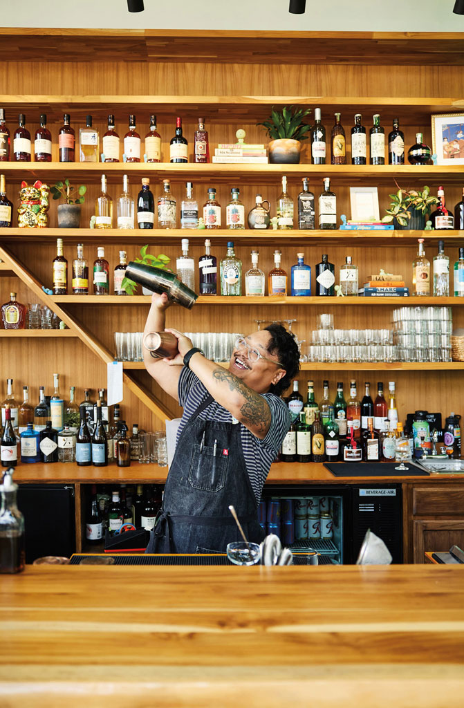 A bartender in glasses and an apron enthusiastically shakes a cocktail shaker behind a wooden bar in the heart of the Chinatown-International District, with shelves full of liquor bottles and glasses gleaming in the background.
