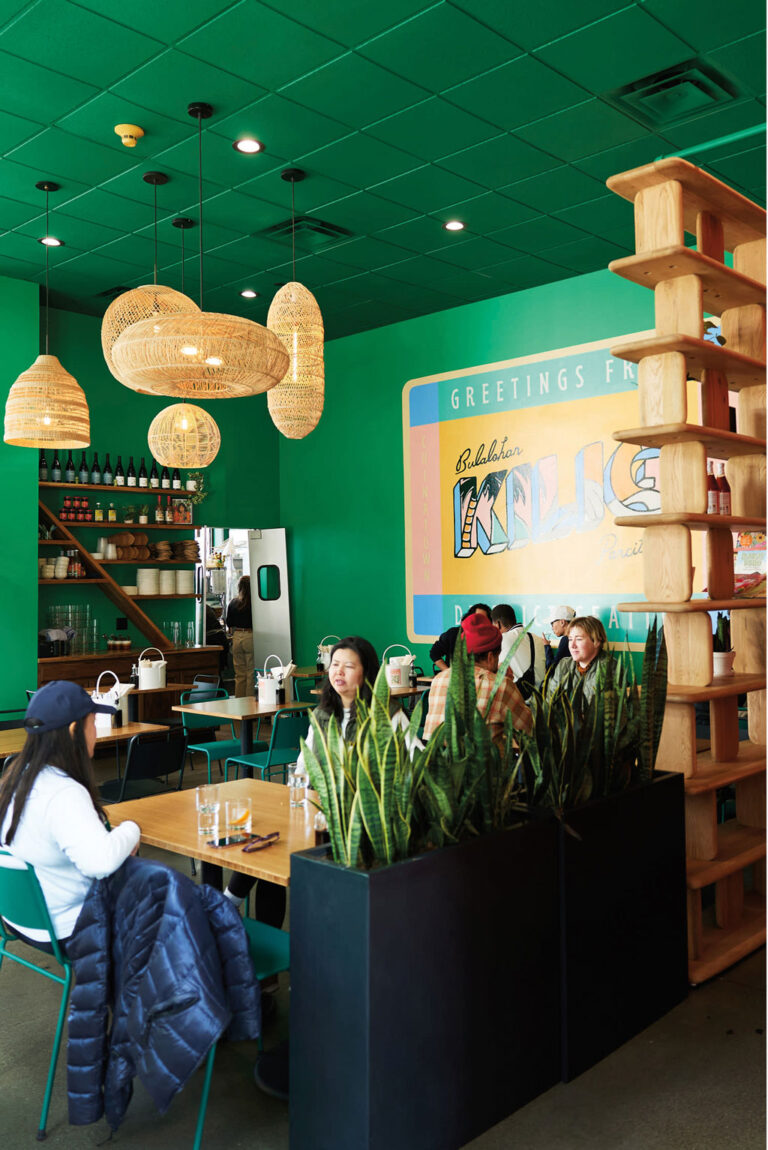 A cozy restaurant in the Chinatown-International District with green walls and ceiling, rattan pendant lights, wooden shelves, and several people at tables. A large mural says "Greetings From Milwaukee," and plants divide the seating area.