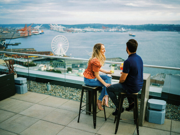 A woman and man sit on barstools at a rooftop patio in a downtown neighborhood of Seattle, overlooking a harbor with a ferris wheel, docks, and water, enjoying drinks and talking.