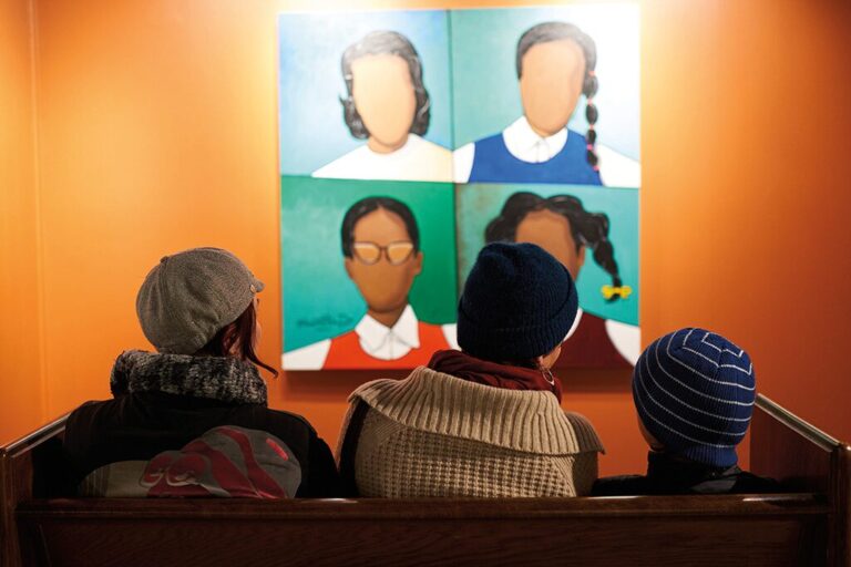 Three people sit on a bench facing a bright orange wall, looking at a painting of four faceless girls with different hairstyles and clothes. The viewers wear winter hats and jackets. The scene feels contemplative.