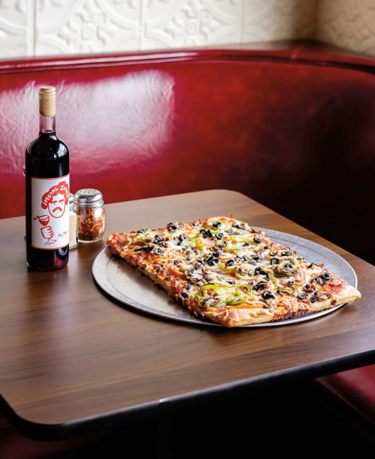 A rectangular pizza with olives, green peppers, and onions on a metal tray sits on a wooden table, next to a bottle of red wine and a jar of crushed red pepper, with a red booth seat in the background.