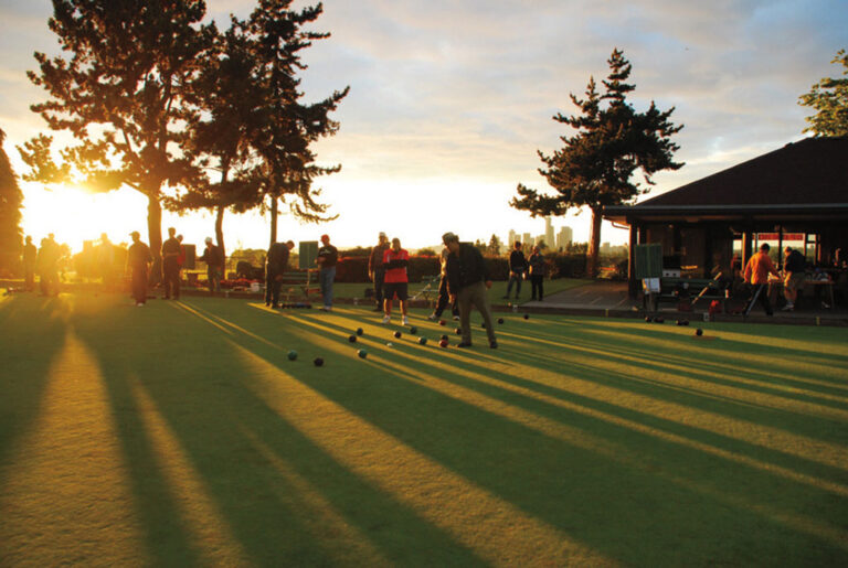 People play lawn bowling on a green field at sunset, casting long shadows. Trees and a building are in the background, with the sun low in the sky, creating a warm, golden light.