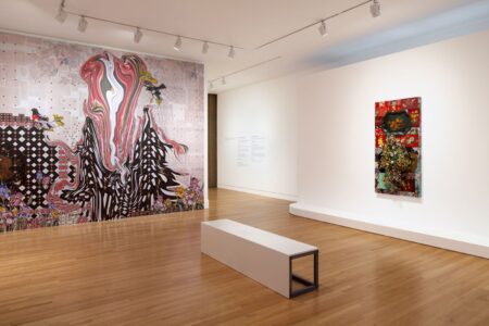 A modern art gallery with wooden floors features a large, colorful mural with abstract patterns and birds on one wall, a rectangular white bench, and a vibrant mixed-media artwork on an adjacent wall.