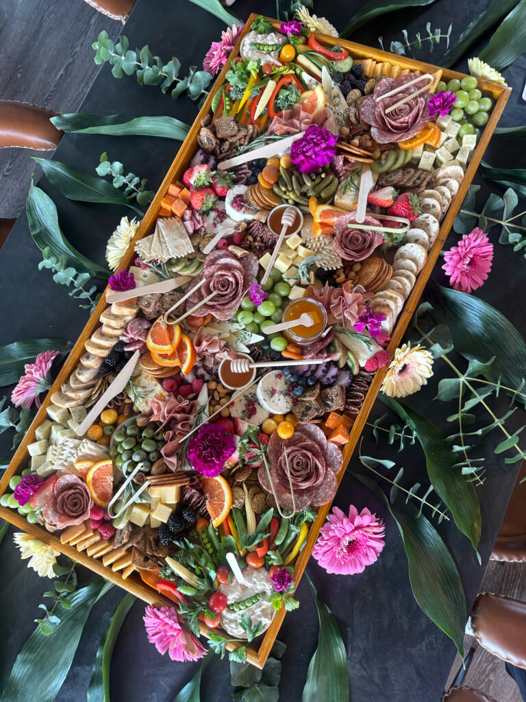A large wooden tray filled with an assorted charcuterie spread, including meats, cheeses, crackers, fruits, nuts, and garnished with flowers. Green leaves and colorful blossoms surround the tray on a dark table.
