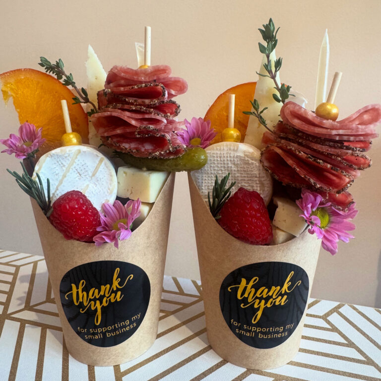 Two brown cups labeled "thank you for supporting my small business" are filled with charcuterie items, including salami, cheese, pickles, raspberries, an orange slice, and pink flowers, arranged artfully.