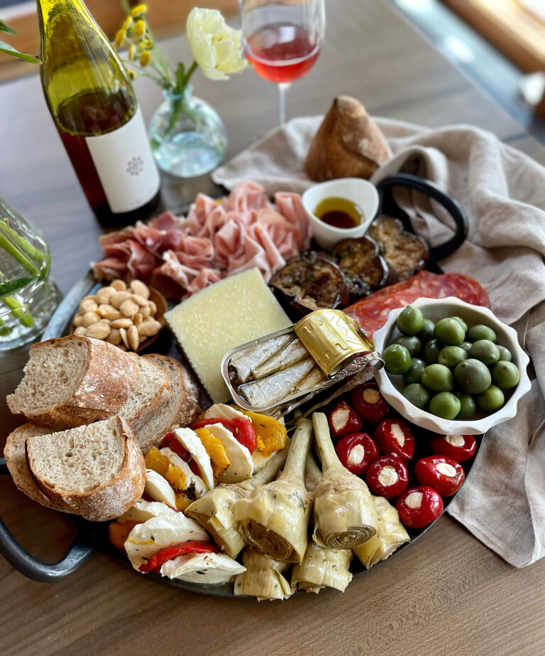A charcuterie platter with bread, cheese, prosciutto, salami, marinated artichokes, grilled vegetables, stuffed peppers, green olives, almonds, and a bottle and glass of rosé wine on a wooden table.