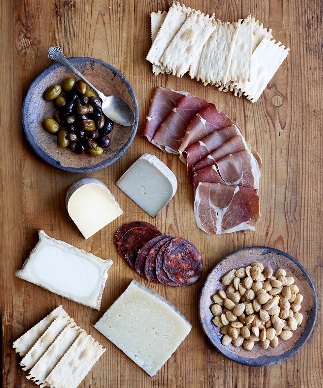A wooden table with an assortment of crackers, sliced cured meats, three types of cheese, marinated olives in a bowl with a spoon, spicy sausage slices, and a small plate of toasted almonds.