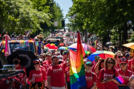 A large crowd marches in a Pride parade on a sunny day, many wearing red shirts and carrying rainbow flags and umbrellas. Decorated floats and trees line the street, with people celebrating diversity and inclusion.