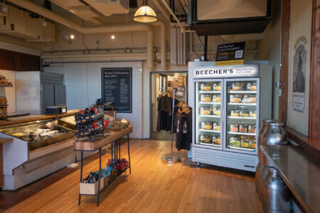 A gourmet cheese shop interior with wooden floors, a refrigerated display filled with cheeses, shelves with products, a counter on the left, and hanging racks of clothing in the back. Large pipes and bright lighting are visible above.