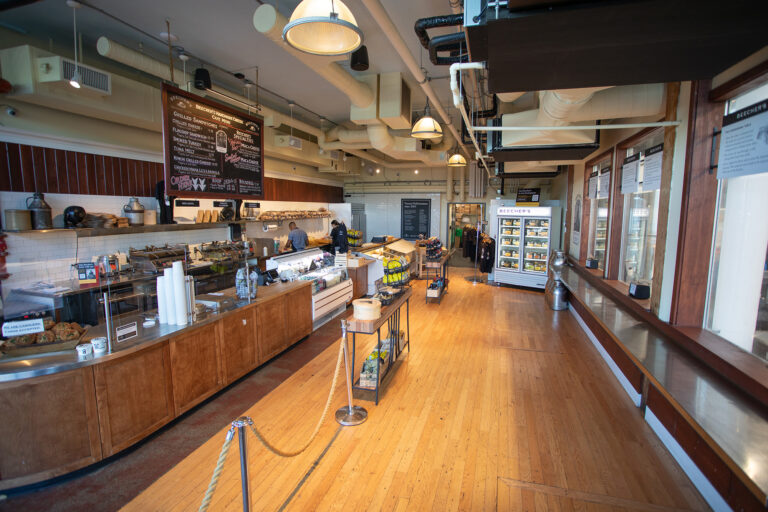 A spacious, well-lit deli with wooden floors, a long counter displaying food items, refrigerated shelves on the right, and a chalkboard menu on the back wall. A few staff members work behind the counter.