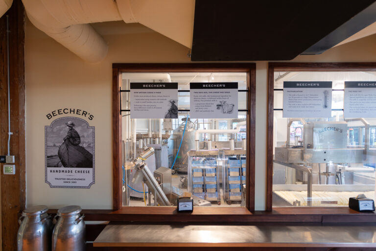 A cheese production area with large metal tanks, equipment, and racks of cheese visible through windows. Signs for Beecher’s Handmade Cheese are displayed above and beside the windows.