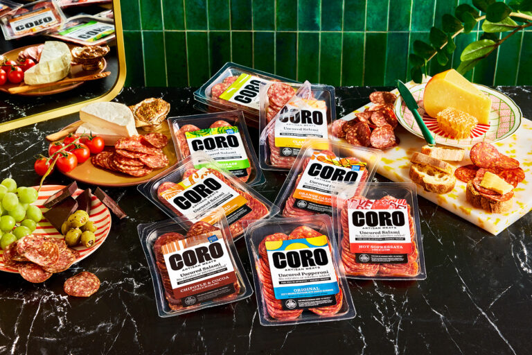 An assortment of Coro brand cured meats in plastic packaging is arranged on a black marble table, surrounded by cheese, grapes, tomatoes, honey, crackers, and charcuterie on colorful plates. A green tiled wall is in the background.