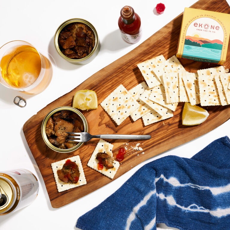 A wooden board with saltine crackers, two opened tins of smoked oysters, lemon wedges, and hot sauce. Some crackers are topped with oysters and sauce. Nearby are beer cans, a glass of beer, and a blue napkin.