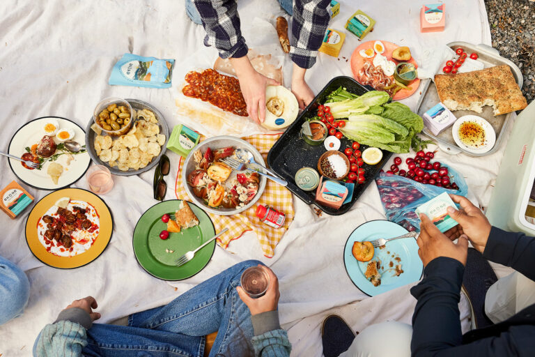 Overhead view of a picnic spread on a white blanket with various plates of food, bread, cheese, olives, fruit, and drinks. Several people are reaching in to serve themselves.