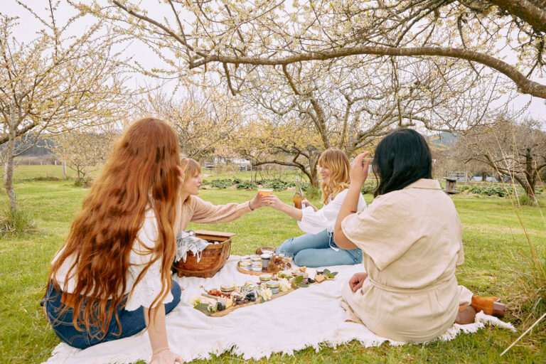 Four women sit on a picnic blanket in an orchard, surrounded by trees. They are smiling and raising drinks in a toast, with snacks and food spread out on the blanket around them. The atmosphere is relaxed and cheerful.