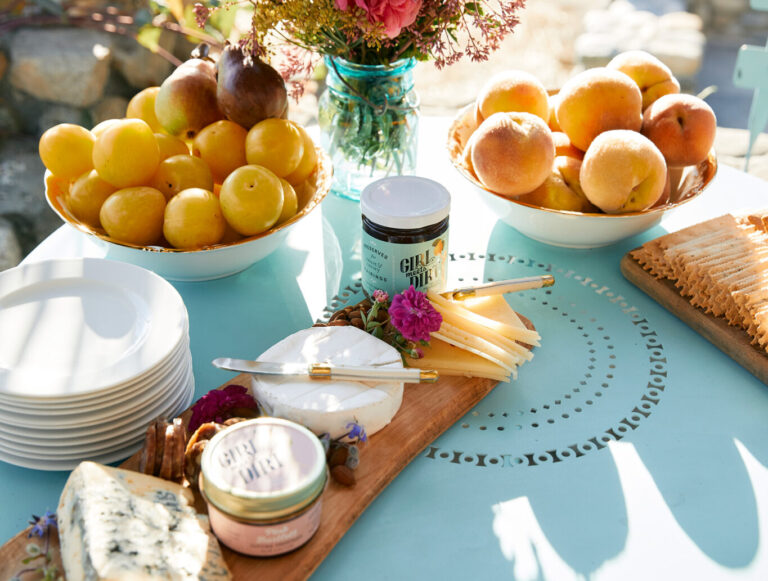 A light blue table set outdoors with bowls of yellow apples and peaches, a vase of flowers, cheese, crackers, and a stack of white plates, bathed in warm sunlight.