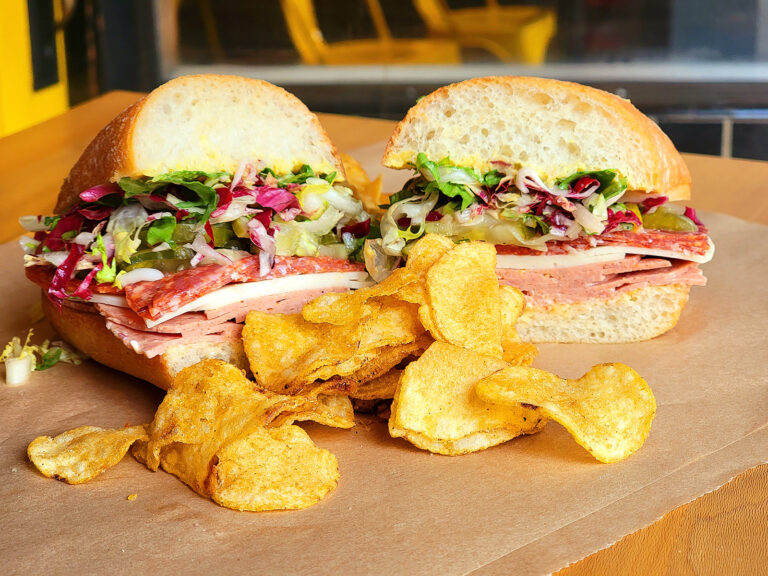 A sandwich filled with cold cuts, lettuce, and greens is cut in half and served on brown paper, with a pile of crispy potato chips in front. Yellow chairs are visible in the blurred background.