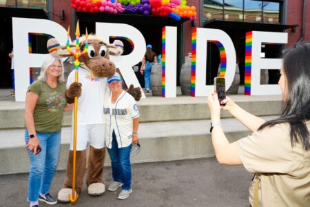 Three people pose with a costumed moose mascot holding a pride flag staff in front of large 