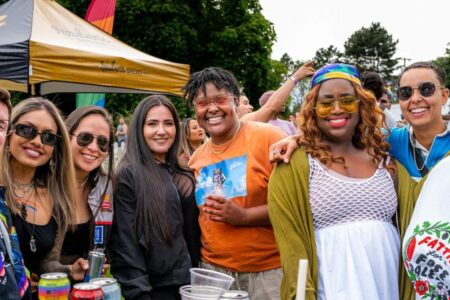 A group of people at an outdoor event smile and pose together. They wear colorful, casual clothing and sunglasses, with tents, trees, and other attendees visible in the background. The atmosphere appears festive and joyful.