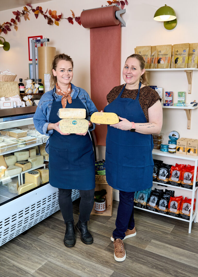 Two women wearing blue aprons stand inside a cheese shop, smiling and holding large blocks of cheese. Behind them are shelves with packaged goods and a refrigerated display case filled with cheese.