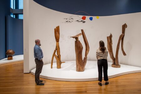 Two people stand in a gallery observing wooden abstract sculptures and a hanging mobile with red, yellow, and blue shapes, displayed on a curved white platform against a blue wall.