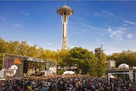 A large crowd gathers at an outdoor music festival near a colorful stage, with the Space Needle towering in the background and trees surrounding the venue under a clear blue sky.