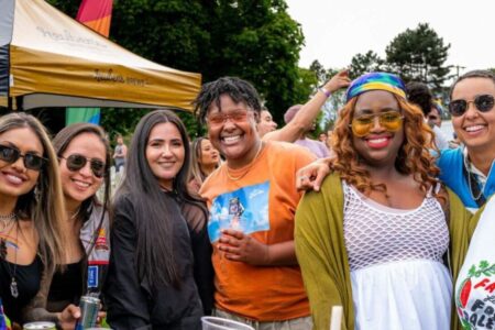 A group of six people stand close together outdoors at a festival, smiling at the camera. They wear colorful, casual clothes and sunglasses, with tents and trees visible in the background.