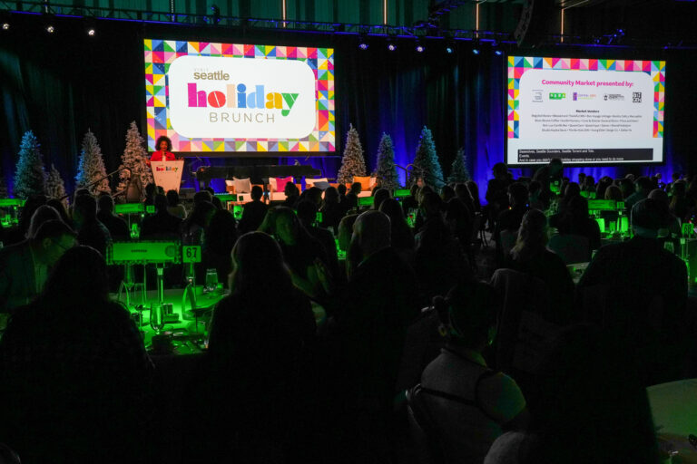 A large crowd attends the Seattle Holiday Brunch in a dimly lit room with green lighting, colorful signage, and Christmas trees on stage. Two large screens display event information and presentations.