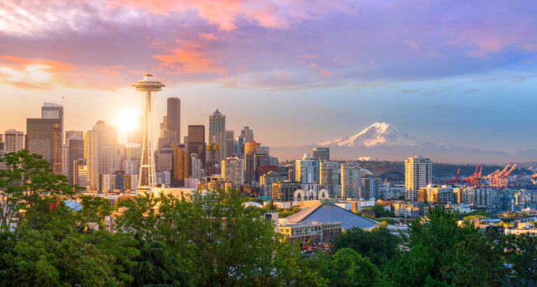 Seattle skyline at sunset with the Space Needle in the foreground, downtown skyscrapers, and Mount Rainier visible in the background under a colorful sky. Lush green trees are in the foreground.