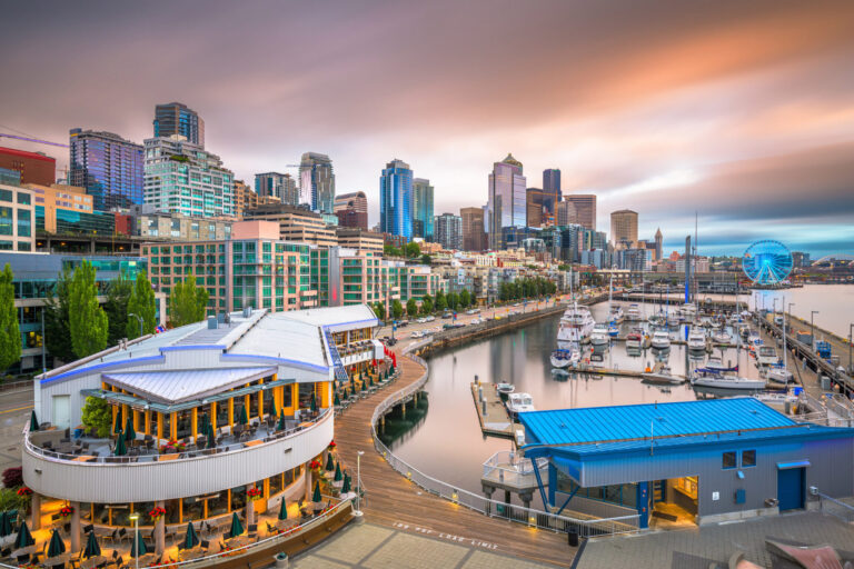 A waterfront view of downtown Seattle at sunset, featuring a marina with boats, waterfront restaurants, modern buildings, and the Seattle Great Wheel near the edge of the bay.