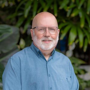 A smiling bald man with glasses and a white beard, wearing a light blue button-up shirt, stands in front of lush green foliage.