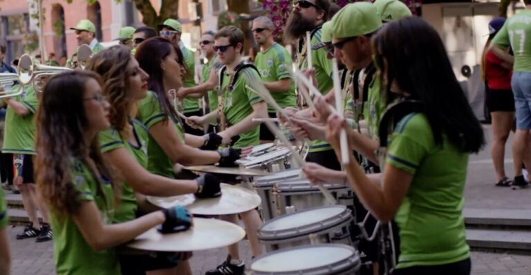 A group of people in green shirts play drums and brass instruments in an outdoor setting, performing as a band or drumline. Trees and buildings are visible in the background.