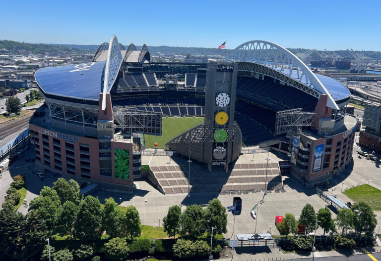 Aerial view of Lumen Field, a premier venue for Seattle sports, with its partially open roof, arched supports, and surrounding trees. The empty field and stands are visible under a clear blue sky.
