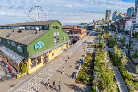 A sunny day at Seattle’s waterfront shows Pier 56 with Elliot’s Oyster House, people walking along the boardwalk, green landscaping, and the Seattle Great Wheel with downtown buildings in the background.