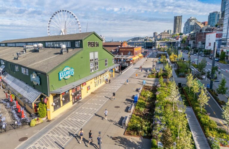 A sunny day at Seattle’s waterfront shows Pier 56 with Elliot’s Oyster House, people walking along the boardwalk, green landscaping, and the Seattle Great Wheel with downtown buildings in the background.