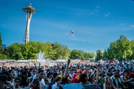 A large crowd gathers at an outdoor festival near a fountain, with the Space Needle and green trees in the background under a clear blue sky—a perfect day for enjoying Seattle events.