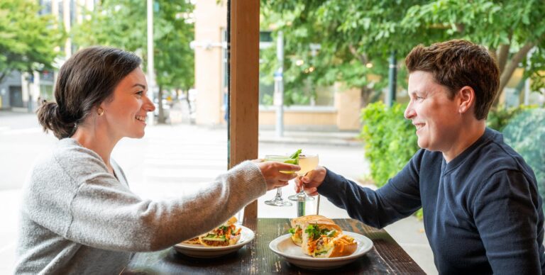 Two people smile and clink cocktails across a wooden table at a restaurant, each with a plate of food in front of them. Large windows reveal green trees and an urban street outside.