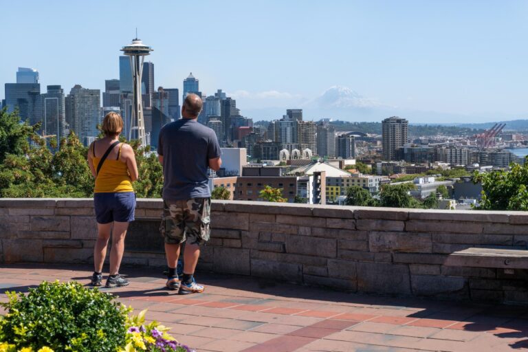 Two people stand at a viewpoint overlooking the Seattle skyline, with the Space Needle visible and mountains in the distance on a clear, sunny day. Lush greenery and flowers frame the foreground.